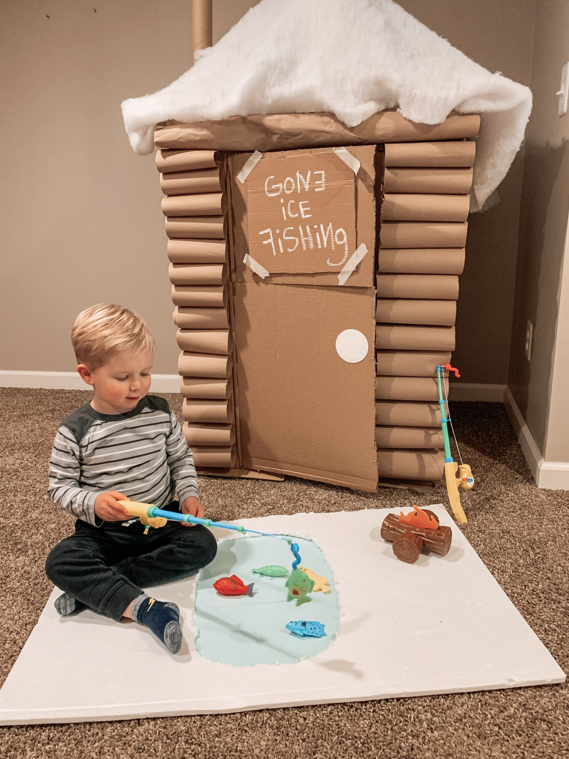 Kid playing with a repurposed cardboard box ice fishing cabin and pretending to fish on packing foam ice